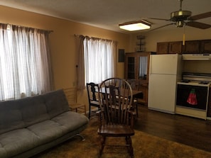 A living room with a sofa, a dining table with chairs, a kitchen area with a refrigerator and stove, and a ceiling fan. - The Pines at Mayhill (Mayhill)