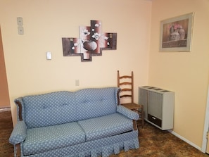 A room with a blue patterned sofa, a wooden chair, a wall-mounted air conditioner, and a framed artwork. - The Pines at Mayhill (Mayhill)