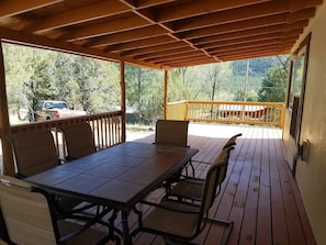 Eine überdachte Terrasse mit Tisch und Stühlen, einem Hollerleger und Blick auf ein Fahrzeug und Bäume im Hintergrund.