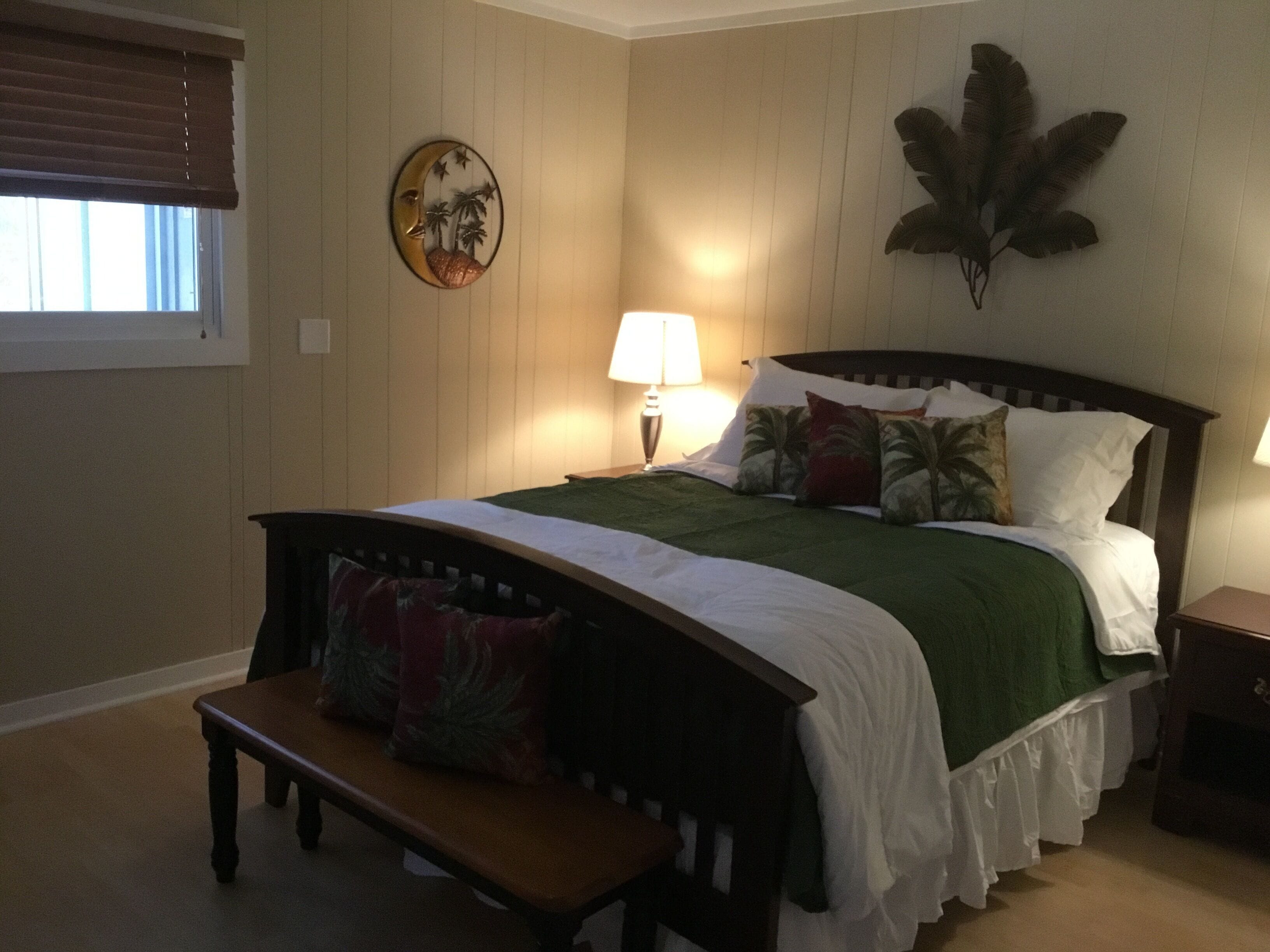 Een slaapkamer met een bed, een nachtkastje, een bankje, een wandklok en een raam met blinds.