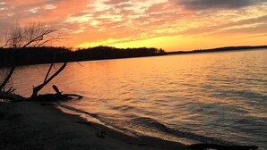 Um pôr do sol sobre águas calmas, com uma orla arenosa e árvores sem folhas.