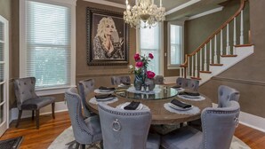 A dining room with a round glass table, grey upholstered chairs, a chandelier, a framed portrait, and a staircase in the background. - Amazing Home & Area - Close to Downtown Nashville! Family & Group Friendly! (Nashville)