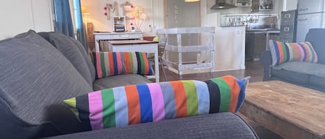 A living room with a grey sofa, colorful striped pillows, a wooden coffee table, and a kitchen in the background.