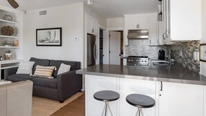 A modern kitchen with white cabinetry, a stainless steel island with stools, a built-in oven, and a backsplash with mosaic tiles. - Playa One Bedroom (253) (Carpinteria)