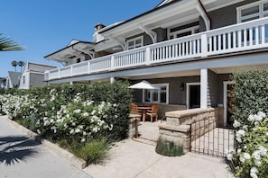 Une maison à deux étages avec un balcon, une terrasse avec une table et des chaises, et des arbustes à fleurs blanches.