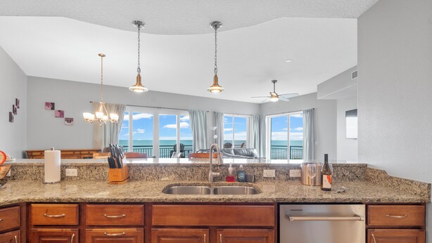 A kitchen with granite countertops, wooden cabinets, a stainless steel sink, and a dishwasher. There are pendant lights and a ceiling fan. - Modern Penthouse with Direct Ocean Front Views! (Cocoa Beach)