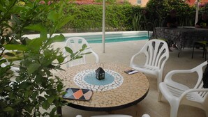 A covered outdoor area with a pool, white plastic chairs, and a table with a mosaic tile design.
