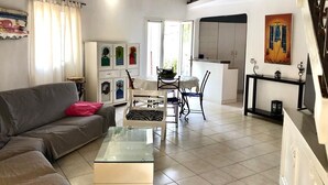 A living room with a sofa, a glass coffee table, and a dining area with chairs and a table. There is a kitchen area with cabinets and a balcony with a railing.