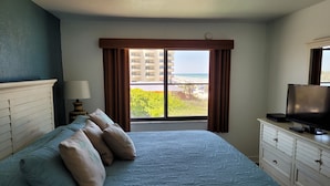 Hotelkamer met een bed, een televisie, een dressoir en een raam met uitzicht op een gebouw en een strand.