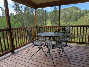 En overdækket terrasse med bord og stole, omgivet af et trægelænder, med udsigt over en skovklædt bakke.