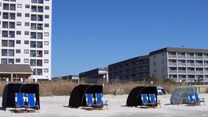 Ein Strand mit mehreren Liegestühlen und Sonnenschirmen, ein hohes Gebäude mit zahlreichen Balkonen und ein weiteres Gebäude mit flachem Dach.