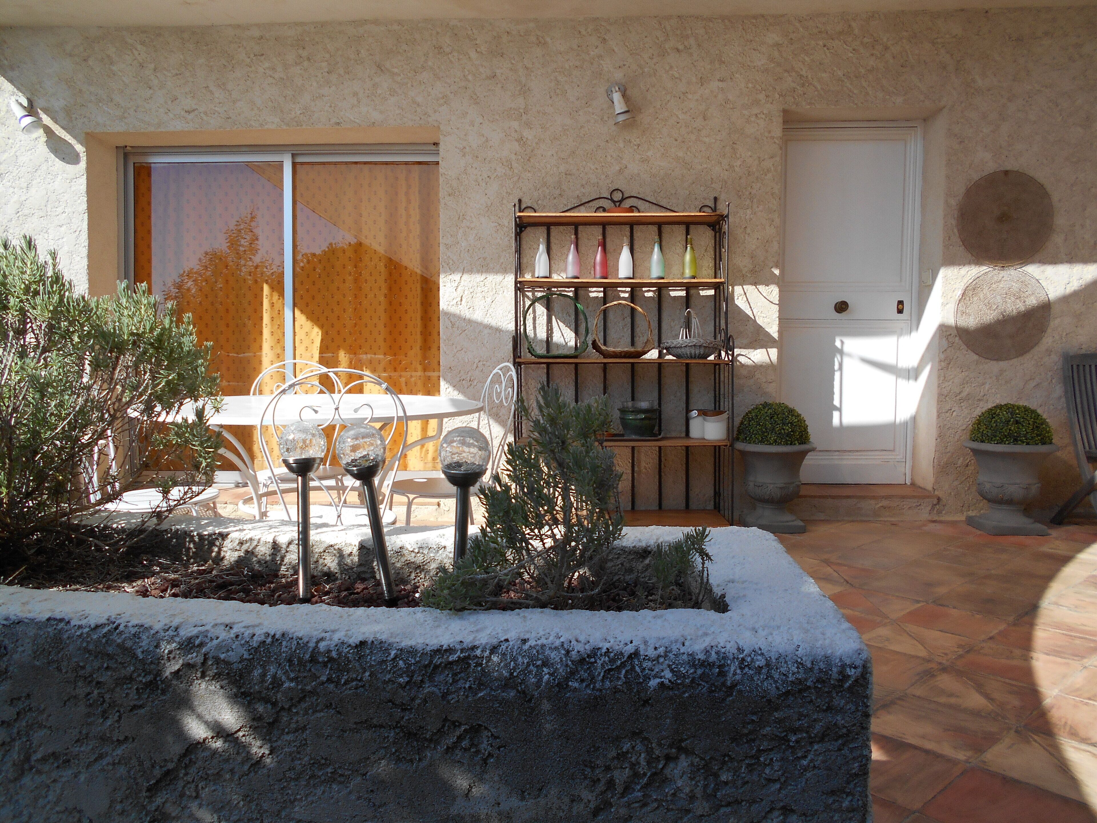 A patio with a white table and chairs, a metal shelf with bottles, and potted plants.