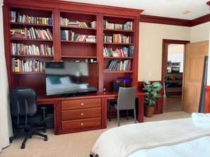 A room with a large wooden bookshelf filled with books, a flat-screen TV, a desk with a chair, a potted plant, and a bed.
