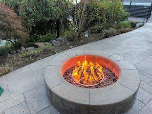A circular stone fire pit with flames, surrounded by a paved walkway and greenery.