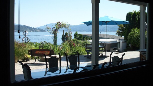 A view through a window of a patio area with a hot tub, outdoor seating, and a view of a lake and mountains. - 3 bed Lake Sammamish Waterfront + Peloton Bike (Bellevue)