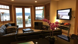 A living room with a sofa, ottoman, and a TV mounted on the wall. There are windows with blinds, a plant, and a shelf with dumbbells. - 3 bed Lake Sammamish Waterfront + Peloton Bike (Bellevue)