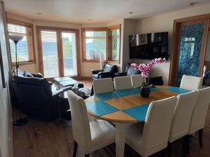 A modern living room with a round table, comfortable seating, a flat-screen TV, and large windows with blinds.