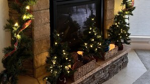 Une cheminée avec un manteau en pierre, décorée pour Noël avec des guirlandes lumineuses, une guirlande à carreaux et de petits sapins.