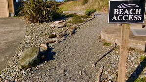 Un chemin de gravier mène à un panneau indiquant l’accès à la plage, le tout étant entouré de végétation naturelle et d’un bâtiment.