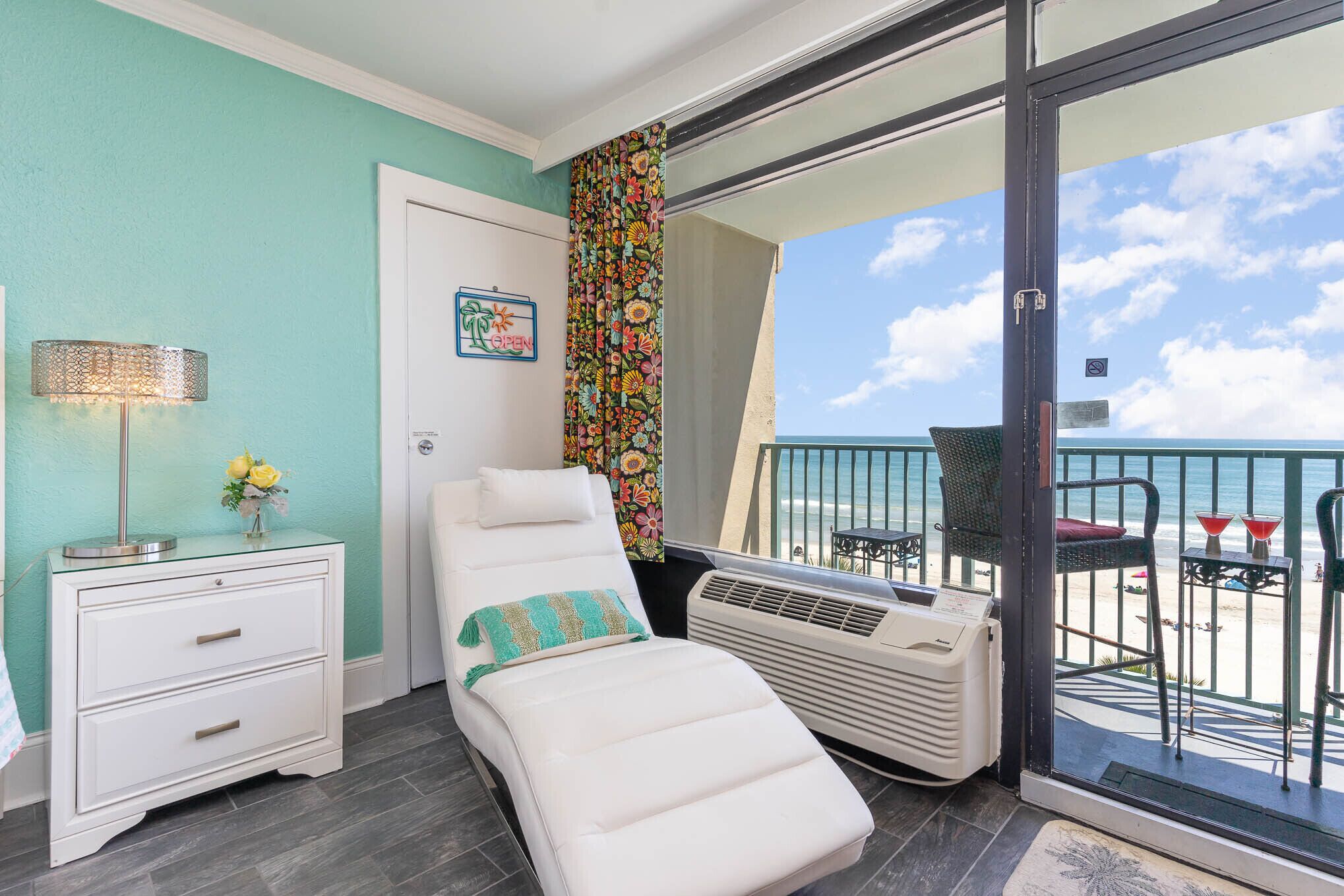 A balcony with a white sun lounger, a small table with a lamp, and a view of the beach.