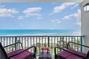 A balcony with wicker chairs and a table, overlooking a beach with people and umbrellas.