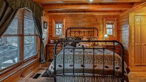 A wooden cabin bedroom with a metal bed frame, a window with blinds, and a wooden door. - 5 BR Log Home - 5 Min/2.7miles to TIEC - with a Hot Tub on 18 acres "Creekside"  (Rutherfordton)