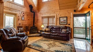 A cozy log cabin interior with a leather sofa, a fireplace, and a large window. - 5 BR Log Home - 5 Min/2.7miles to TIEC - with a Hot Tub on 18 acres "Creekside"  (Rutherfordton)