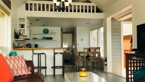 Un espacio habitable amplio con un sofá de cuero marrón, una mesa de centro de vidrio y un comedor con sillas de madera, con una cocina al fondo.