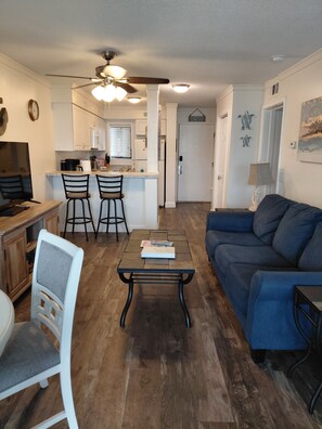 A living room with a blue sofa, a coffee table, and a TV. There is a kitchen area with a refrigerator, stove, and bar stools.