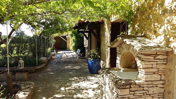 Un sendero de piedra conduce a un área de estar rústica al aire libre, con una pérgola arriba, rodeada de abundante vegetación.