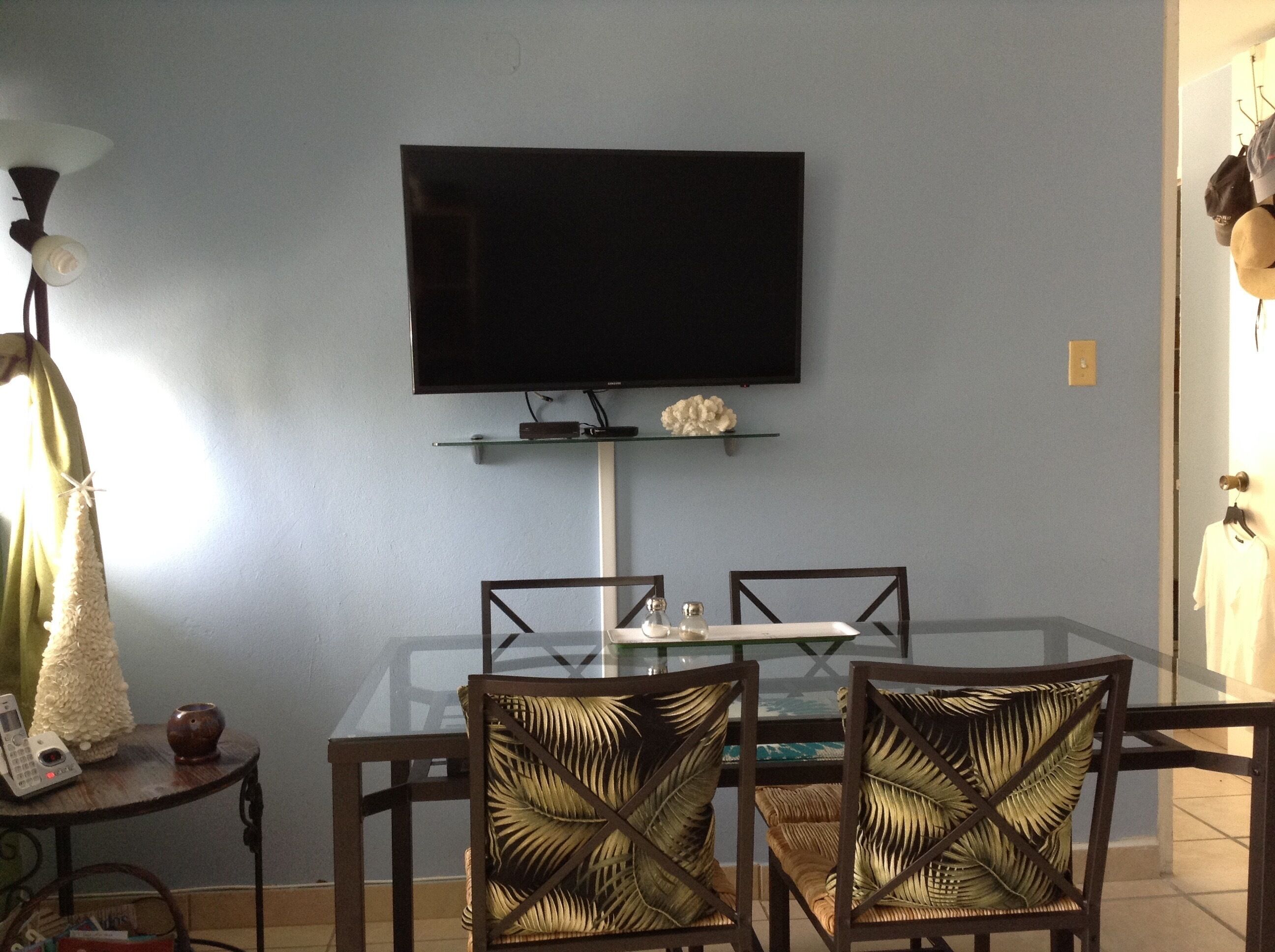 Un comedor con una mesa de vidrio, cuatro sillas de diseño trenzado, un televisor de pantalla plana montado en la pared y un pequeño estante debajo del televisor.