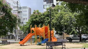 Un parque infantil con toboganes y estructuras para escalar, bancos y un estacionamiento con una camioneta.