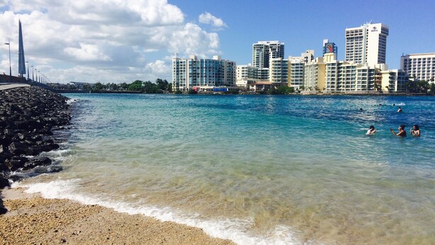 Una playa con agua turquesa cristalina, una costa rocosa y un paisaje urbano con rascacielos al fondo.