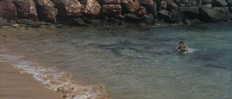 En klippefyldt bro med mennesker, der sidder og står, med udsigt over en strand med klart vand.