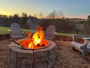 A circular stone fire pit with two Adirondack chairs facing it, set in a gravel area with a view of trees and hills in the background. - Blue Ridge, Tye River, C-Ville, Crabtree, Wintergreen, Sweet Briar, Farm (Arrington)