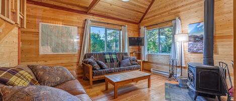 A cozy cabin interior with wooden walls and floors, a sofa, a coffee table, a fireplace, and large windows.