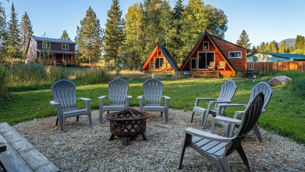 A cabin with a deck, surrounded by Adirondack chairs and a fire pit, set in a forested area.