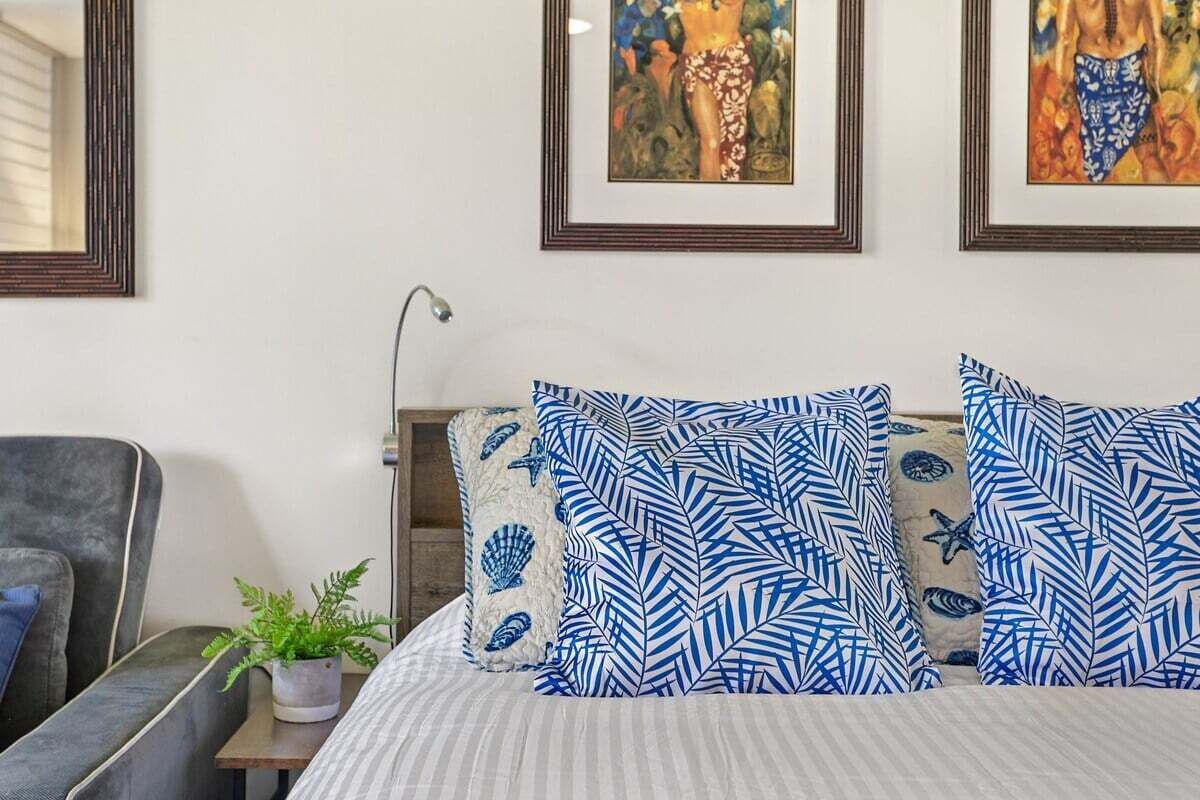 Un dormitorio con una cama, almohadas, una mesita de noche, una lámpara, una silla y una pared decorada con cuadros.