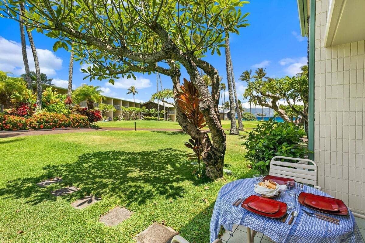 A dining area with a table set for two, a large tree, and a view of a pool and building in the background.