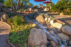 Relax in a serene garden oasis with a natural water feature, surrounded by lush greenery and large rocks.