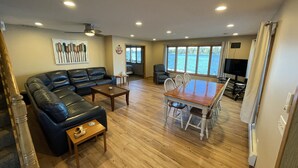 A spacious living room with a black leather sofa, a wooden coffee table, and a dining table with chairs. There is a large window offering a view of a lake. - Lakefront Family Getaway! Pontoon Rentals-Kayak-Pedal Boat-Fire Pit-Sandy Beach (Lindstrom)
