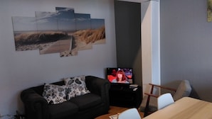 Un salon avec un canapé gris foncé, une table basse et un téléviseur posé sur un meuble. Des œuvres d’art ornent le mur au-dessus du canapé, et une petite table se trouve à proximité, avec une télécommande dessus.