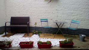 Une terrasse avec un banc, deux chaises, une petite table et des plantes en pot, le tout adossé à un mur de briques blanches.