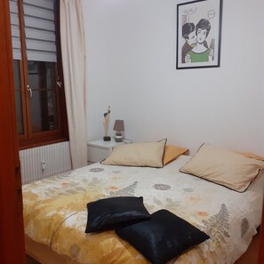 A bedroom with a bed, pillows, a nightstand, a lamp, a framed picture, and a window with curtains. - Apartment with parking in the medieval city, 100 m from the quay, shops. (Saint-Valery-Sur-Somme)