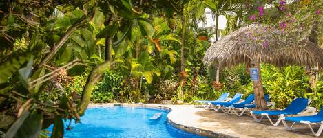 A swimming pool with clear blue water, surrounded by lush greenery and a thatched-roof structure.