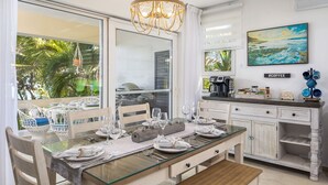 A dining area with a glass table, white chairs, and a chandelier. There is a view of palm trees through the window. - Cabarete - on Kite Beach - Luxury Beachfront Condo w Gardens, Pool & Internet   (caberate)