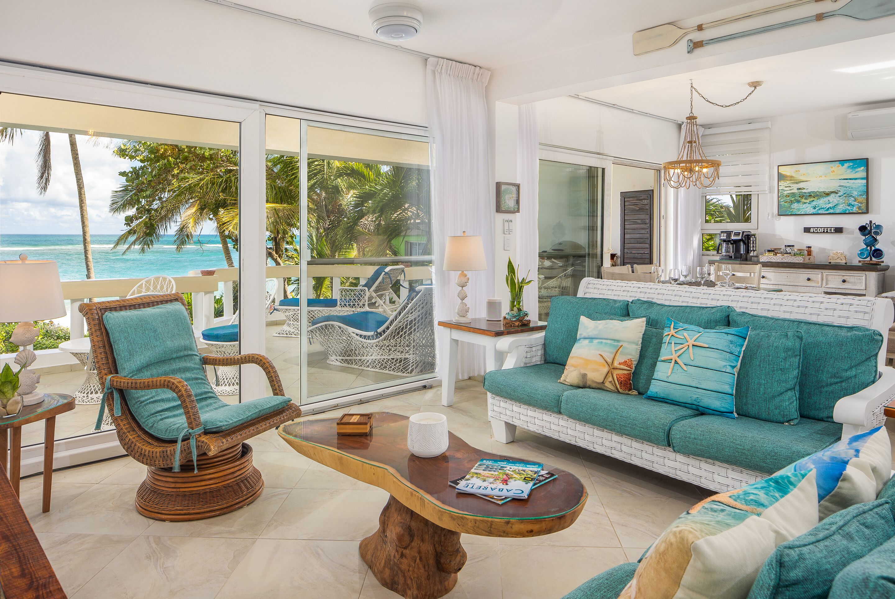 A spacious living room with a view of the ocean, a cozy seating area with a wicker chair, a teal sofa with starfish pillows, and a wooden coffee table with magazines.
