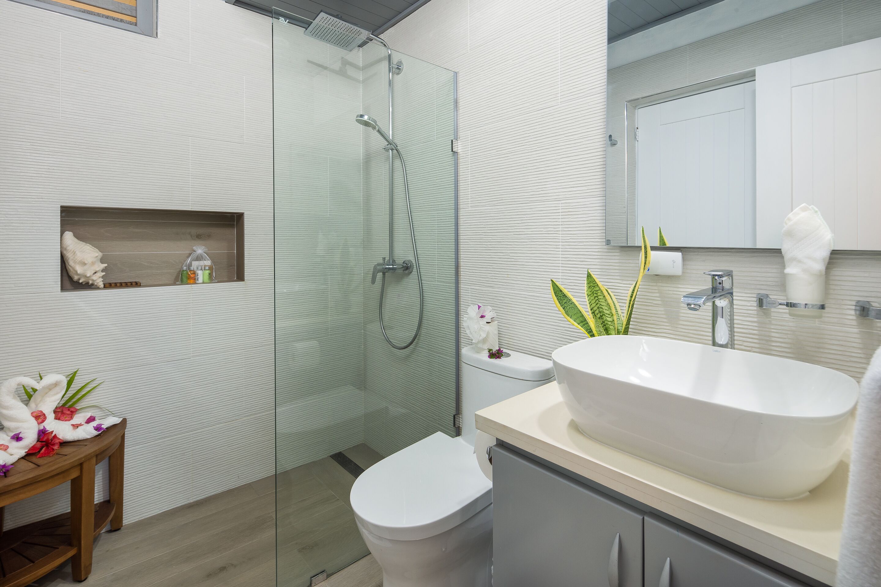 A modern bathroom with a glass-enclosed shower, a white toilet, a wooden vanity with a large sink, and a small shelf with toiletries.