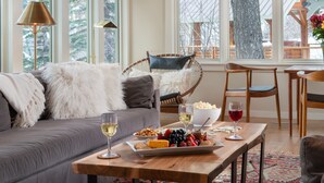 A cozy living room with a grey sofa, fluffy pillows, a wooden coffee table with snacks and drinks, and a view of a snowy outdoor deck. - Ski-In Family Lodge w/ Hot Tub, Game Room, Sauna & Designer Interiors (Teton Village)
