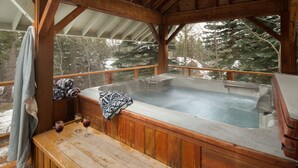 A wooden hot tub with a bench and towels, surrounded by a wooden deck and glass railings, overlooking a snowy landscape. - Ski-In Family Lodge w/ Hot Tub, Game Room, Sauna & Designer Interiors (Teton Village)